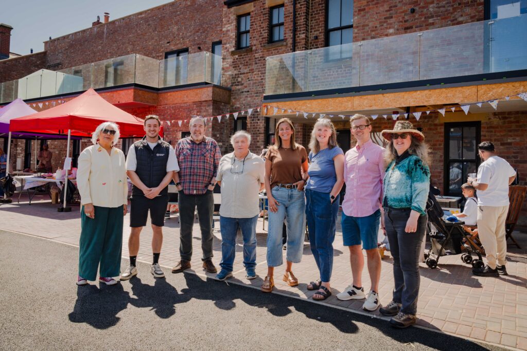 Group of adults standing together outdoors in front of a brick building with tents and bunting, posing for a photo at a community event.