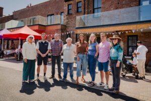 Old Police Station Whitley Bay | H Social | H Social Group of adults standing together outdoors in front of a brick building with tents and bunting, posing for a photo at a community event.